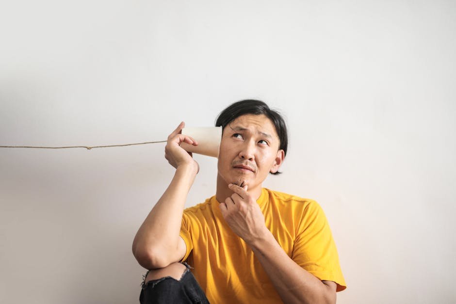 Puzzled Asian male in bright t shirt looking up while listening to answer in paper cup phone on white background